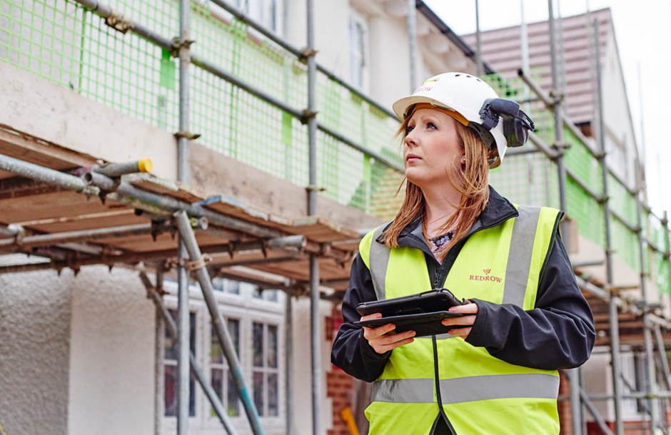 Female professional construction worker in hi-vis jacket and hard hat holding a tablet looking at a building with scaffolding 